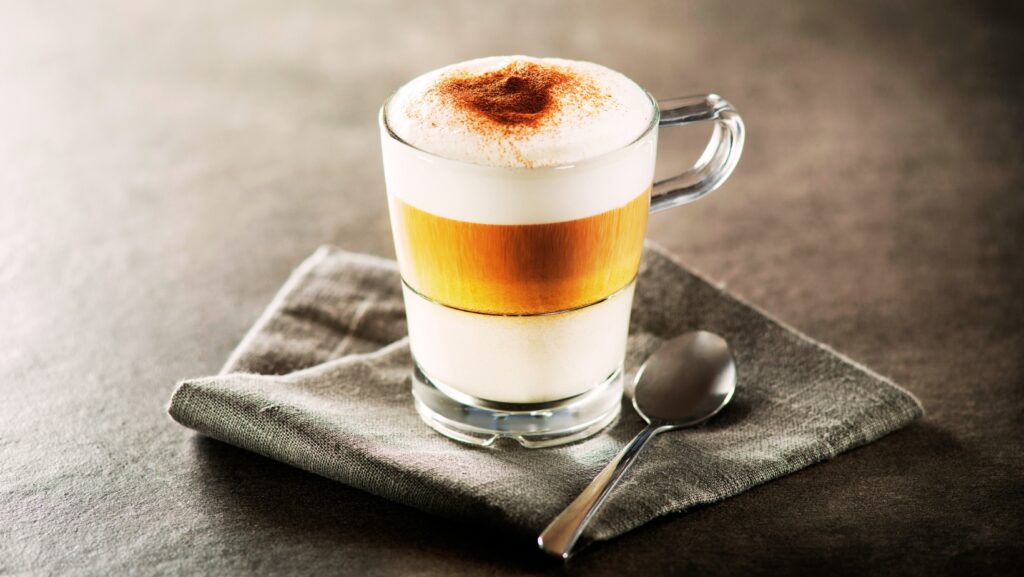 Latte macchiato w wysokiej szklance z uchwytem, z widocznymi warstwami mleka i espresso, posypane kakao, podane na szarej serwetce z łyżeczką, kawa włoska, restauracja Pirat w Unieściu